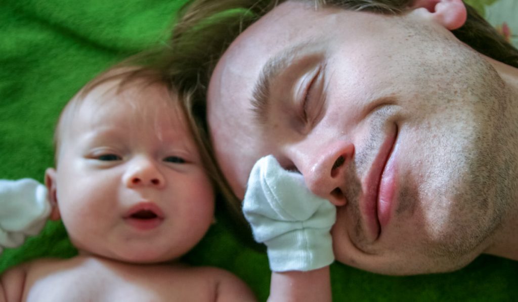Father and baby in mittens lie face to face on a green bedspread.