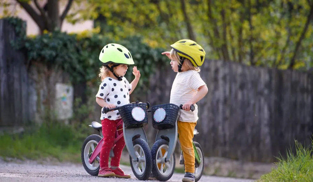 Small children boy and girl with helmets and balance bikes outdoors playing
