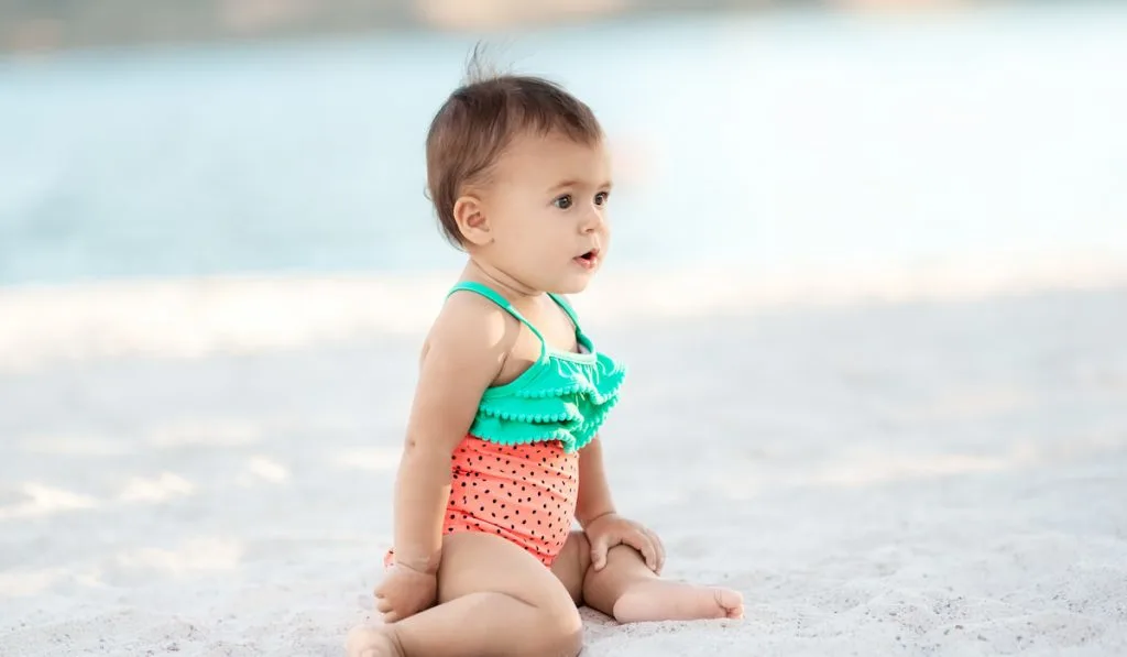 baby girl sitting on sandy beach wearing swimsuit 