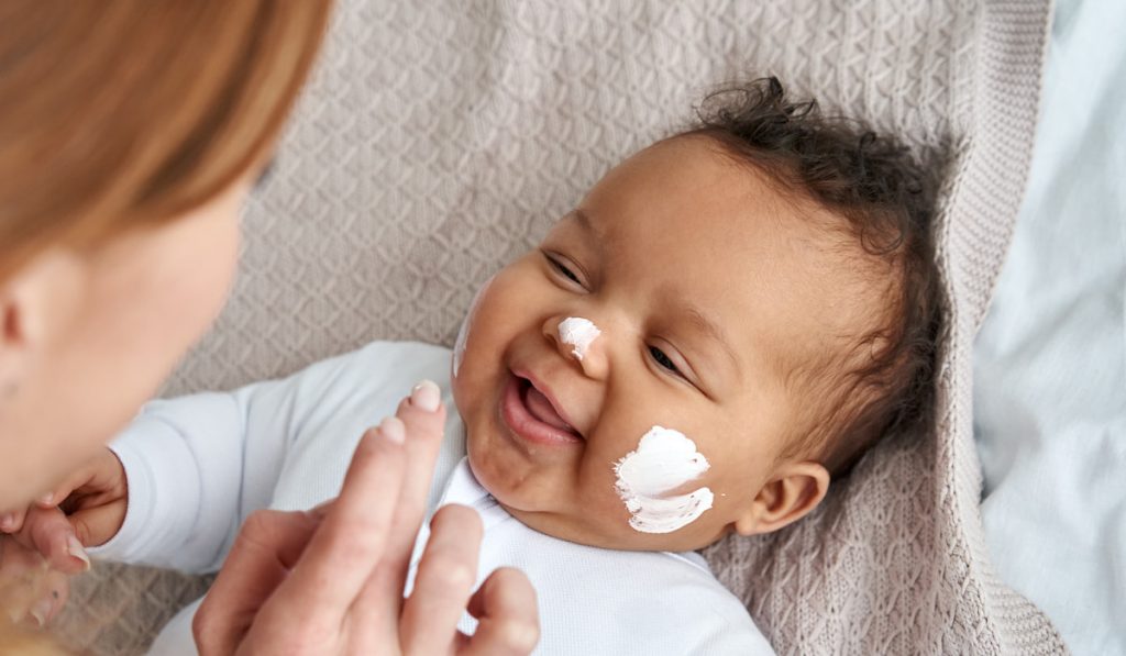 mother applying skin care cream on cute funny baby face.