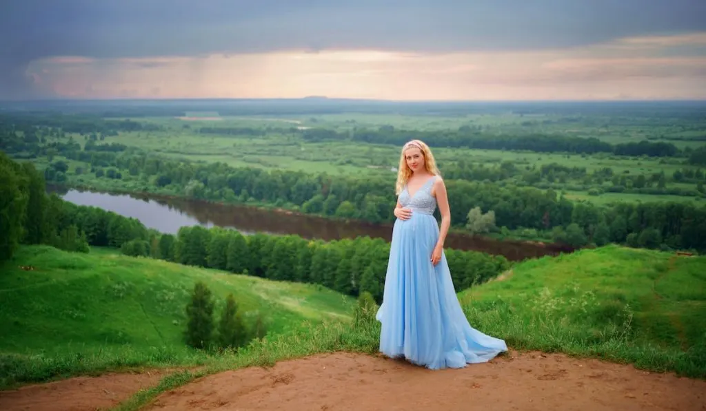 pregnant woman in blue dress, green hills, river and sky