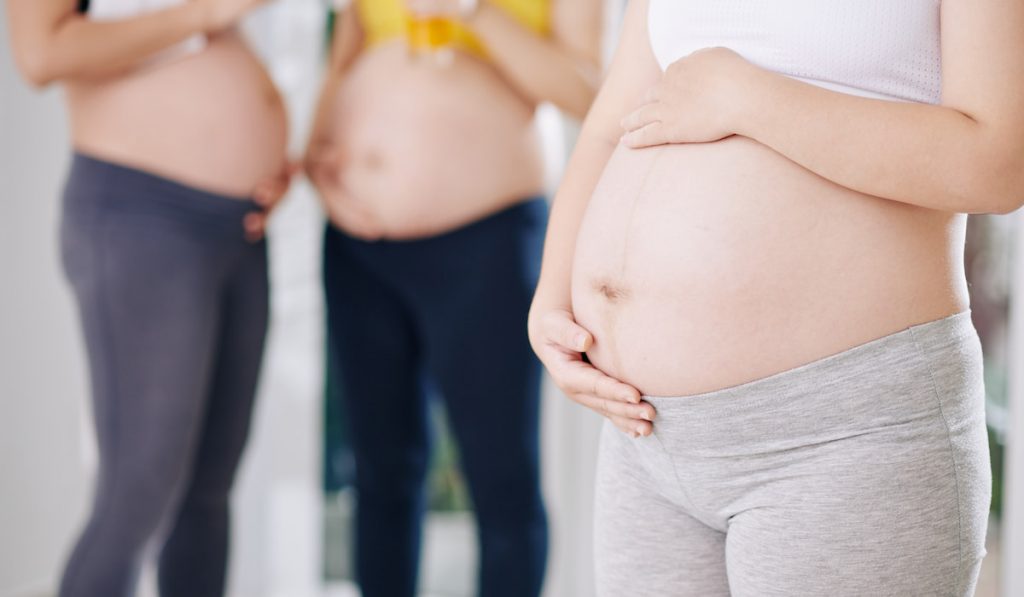 Cropped image of pregnant woman in crop top and soft leggings touching her big belly

