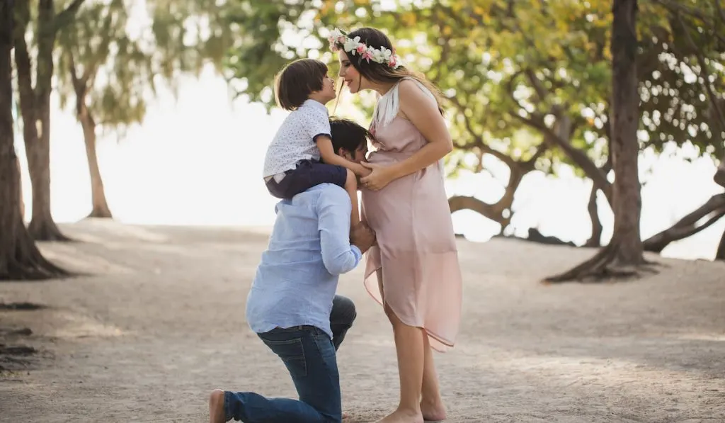 Dad kisses his pregnant wife belly while holds her son on her husbands shoulder