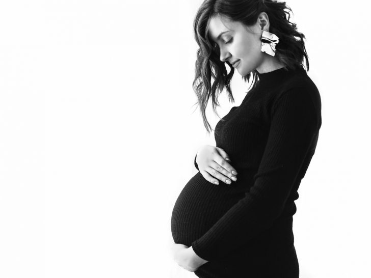 Happy young pregnant woman in stylish black dress holding belly bump and posing
