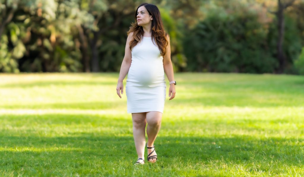 Portrait of a pregnant woman walking in a park with a distracted expression.
