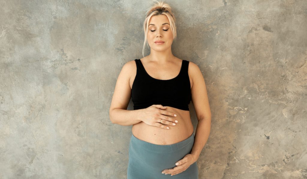 pregnant woman standing near the wall wearing black top and leggings, touching her belly
