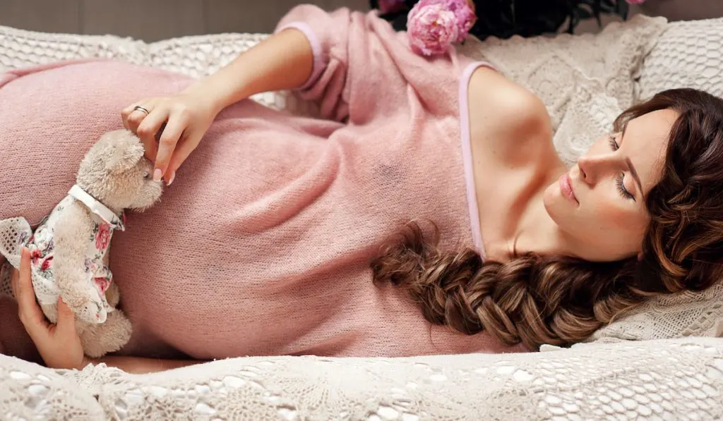 studio portrait of pregnant woman, pink dress, looking in camera, close up, with bear, teddybear, lying on sofa
